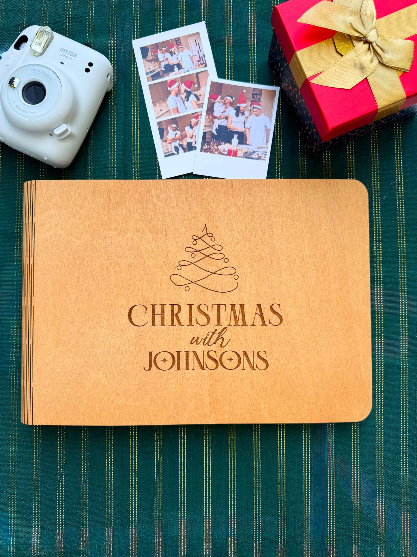 Brown photo album with 'Christmas with Johnsons' engraving, white camera, red gift box, and photos on a green striped background.